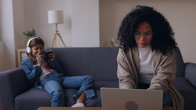 Young woman working on laptop sitting on sofa at home with her daughter playing video games on smartphone. Family conciliation concept.