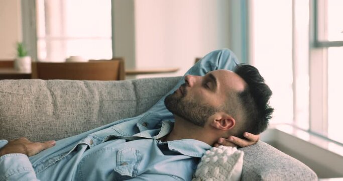 Dreamy peaceful young Hispanic guy relaxing lying on comfortable sofa in cozy living room, deep in pleasant thoughts spend carefree leisure alone at home, enjoy rest, take daytime nap feels satisfied