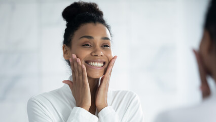 Smiling african American young woman look in mirror in bathroom touch tap healthy face skin after spa beauty procedures, happy biracial female do face massage, apply cream, skincare concept