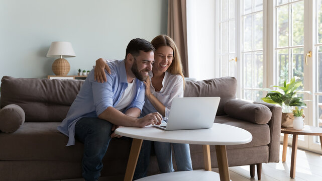 Cheerful millennial family couple using laptop in home living room, looking at monitor with toothy happy smiles, hugging, celebrating good news, success, talking on video call