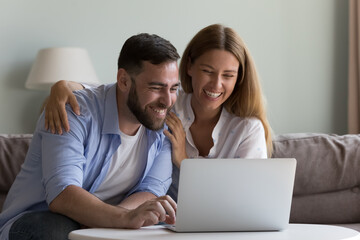 Happy excited millennial husband and wife getting good news from video call on laptop, looking at screen, smiling, laughing, enjoying online communication, using Internet app