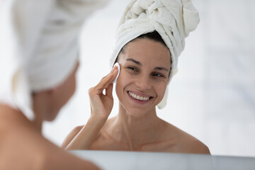 Smiling girl after shower look in mirror cleanse clean face with cotton sponge using lotion or...