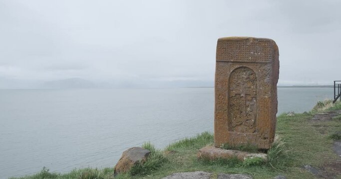 Capture the tranquil beauty of a stone slab adorned with a cross, enveloped by moss and lichen, set against the backdrop of Lake Sevan in Armenia.