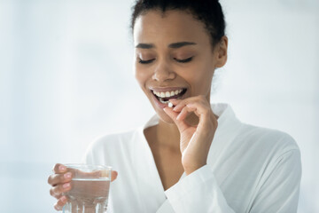 Smiling biracial young woman take daily dose of vitamins or supplements with glass of water in...