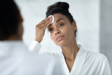 African American woman in bathrobe look in mirror use cotton pad apply toner or lotion on face, biracial young female do beauty procedures for healthy glowing skin in home bathroom, skincare concept