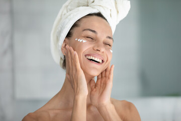 Overjoyed young woman in bathroom after shower apply nourishing moisturizing face cream or mask,...