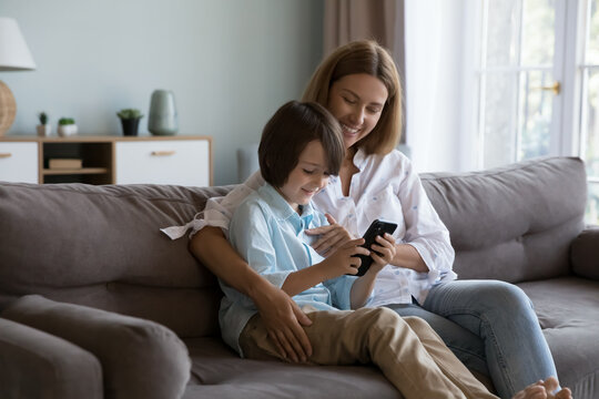 Happy mom teaching curious gen Z son boy to use online learning app on mobile phone, watching child playing virtual video game. Mother and kid sharing mobile phone on home sofa, enjoying communication