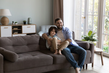 Happy joyful dad and son kid hugging on cozy couch, sitting close together, looking away, watching TV, relaxing, enjoying fatherhood, childhood, family leisure time