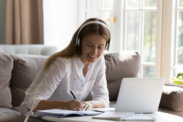 Happy pretty millennial woman using wireless headphones, laptop computer, watching learning webinar, lesson, talking on video call to business client, giving help to customer, writing notes