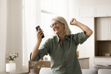 Cheerful excited elderly woman dancing with mobile phone at home, celebrating good dews, win, luck,...
