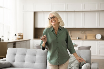 Joyful excited blonde senior woman in glasses enjoying motion, music, party, dancing in living room with closed eyes, laughing. Retired dancer lady relaxing in home interior
