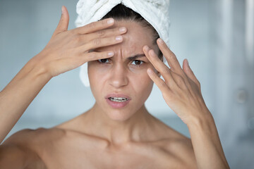 Anxious young Caucasian woman after shower look in mirror in bathroom worried about face wrinkle or...