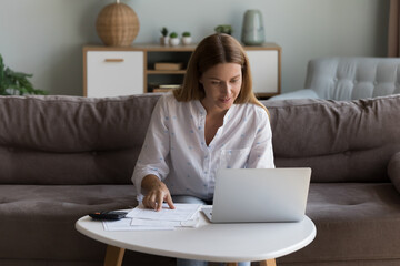 Satisfied pretty millennial laptop user woman checking domestic paper documents, consulting Internet, using banking app for payment, transactions, smiling, sitting at computer on couch at home