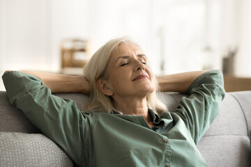 Positive tranquil elderly woman relaxing on couch with closed eyes, smiling, stretching body,...