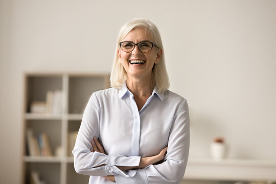 Cheerful Confident Blonde Elderly Business Woman In Elegant Glasses Posing Indoors, Standing With Folded Arms, Looking At Camera, Laughing. Female Professional, Leader Front Portrait