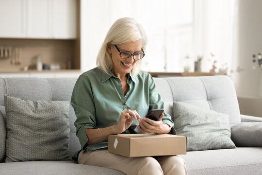 Cheerful old retired consumer woman getting purchase from Internet store, holding paper box on lap, using smartphone, giving positive review, feedback to delivery logistic service, Internet store