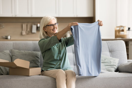 Cheerful blonde elderly woman unpacking parcel from Internet chop at home, taking cloth from carton box, reviewing T-shirt. Online store consumer satisfied with delivery service
