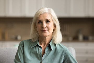Pretty blonde old mature woman looking at camera with serious positive face expression, posing indoors in modern home apartment interior. Video conference call head shot portrait - obrazy, fototapety, plakaty