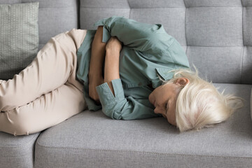 Upset exhausted older woman suffering from strong pain, health problems, serious disease symptoms, feeling stomach ache, resting on home couch with arms folded on belly