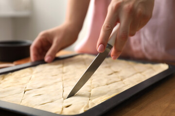 Making delicious baklava. Woman cutting dough in baking pan at wooden table, closeup - obrazy, fototapety, plakaty