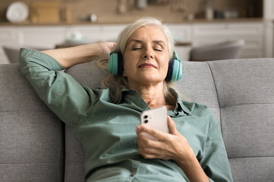 Peaceful Relaxed Senior Lady Enjoying Music, Comfort On Soft Couch At Home, Using Mobile Phones And Big Headphones, Listening To Online Ambient Retro Playlist For Chill, Relaxation