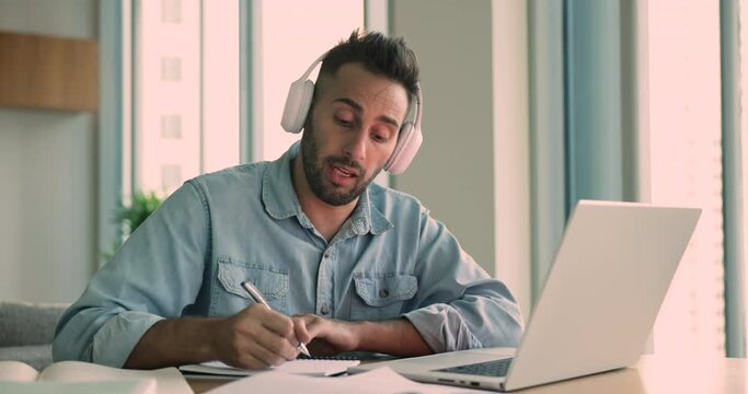 Hispanic young man in headphones sit at desk talk to on-line tutor looking at laptop making notes, studying by video conference call, online training, e-coaching e-learning using modern technologies