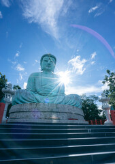 A large statue of Buddha, known as Daibutsu, is seen against a clear blue sky with a bright sunstar shining behind the head