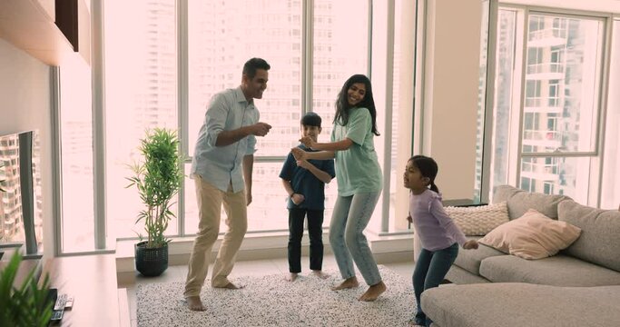 Carefree young Indian parents with little cute son and daughter dancing in modern living room in skyscraper apartment, spend leisure enjoy favourite song, move to music feel happy. Pastime of family