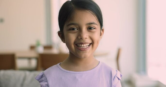 Close up head shot of preschooler Indian girl sitting alone on couch at home smile looking at camera, feel happy and satisfied. Carefree childhood, portrait of candid, beautiful, Alpha generation kid