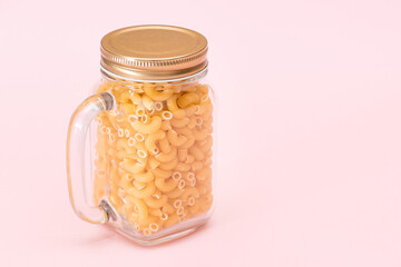 Uncooked Chifferi Rigati Pasta in Glass Jar on Pink Background. Fat and Unhealthy Food. Scattered Classic Dry Macaroni. Italian Culture and Cuisine. Raw Pasta