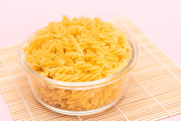Uncooked Fusilli Pasta in Glass Jar on Bambo Mat on Pink Background. Fat and Unhealthy Food. Classic Dry Spiral Macaroni. Italian Culture and Cuisine. Raw Pasta