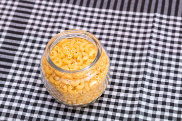 Uncooked Chifferi Rigati Pasta in Glass Jar on Black Checkered Towel. Fat and Unhealthy Food. Scattered Classic Dry Macaroni. Italian Culture and Cuisine. Raw Pasta