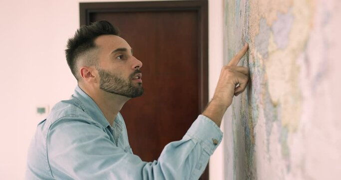 Side view handsome smiling Hispanic man standing next to wall search, review paper world map, pointing on country want to visit, planning next trip. Travel, adventure lover and journey aspirations