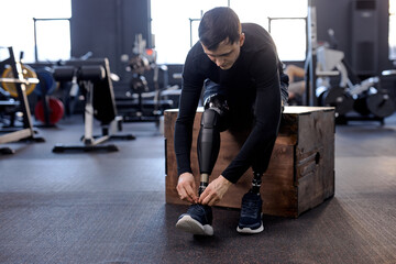 amputee male athlete tying shoelaces sitting on fitness box in gym with modern equipment, full length photo. health and body care - obrazy, fototapety, plakaty