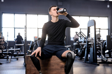 Fototapeta na wymiar A thirsty tired handsome sportsman with artificial leg sitting at sport center drinking refreshment. close up photo, free time, spare time, lifestyle
