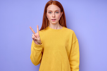 beautiful serious redhead woman in yellow stylish sweater demonstrating letter V sign language symbol for deaf human with blue background. isolated close up portrait