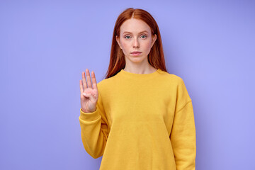 beautiful serious confident redhead woman in yellow stylish sweater demonstrating 4 number sign language symbol for deaf human with blue background. isolated close up portrait