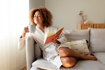 Pensive relaxed Multiracial hispanic woman holding a book, drinking morning coffee sitting on the sofa. - obrazy, fototapety, plakaty