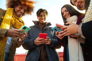 Smiling group of multiracial friends looking at their mobile phone on winter day. Addicted to...