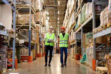 Retail Warehouse full of Shelves with Goods, Two Young Caucasian Male Workers Supervisors in Working Uniform and Helmet Discuss Product Delivery. Men Walking and Having Talk In Warehouse