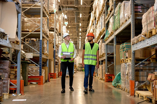 Caucasian engineers in safety helmets and green uniforms walking among shelves with goods in warehouse talking, checking goods. logistic and business export, distribution and inventory concept