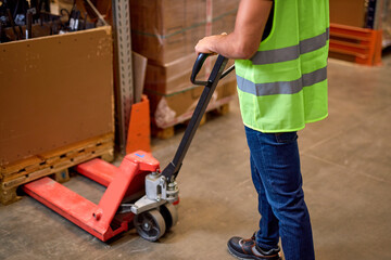 worker in a warehouse uses hand pallet stacker to transport pallets good cargo, cropped unrecognizable male staff in green uniform vest at work place. Hand pallet truck jack in factory warehouse.