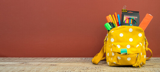 Yellow backpack with white polka dots with different colorful stationery on table. Brown background. Back to school. Banner design.