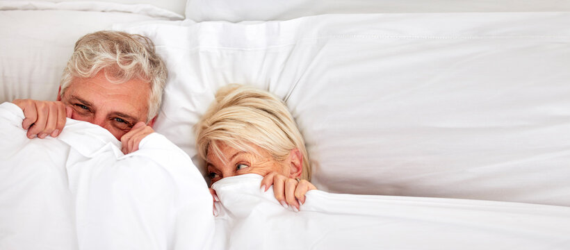 Senior couple, hiding and smile in bed for fun, comfort and bonding for love, silly and goofy together. Elderly people, marriage and sex in bedroom in retirement, romance and happiness at home - Powered by Adobe