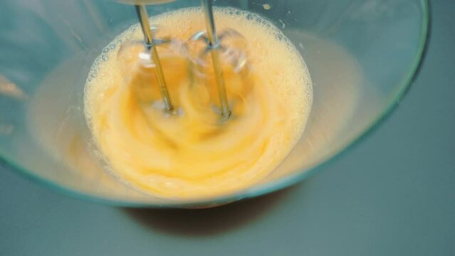 Woman beating eggs with a mixer in a deep glass bowl. Close-up of a table. Cooking in the kitchen