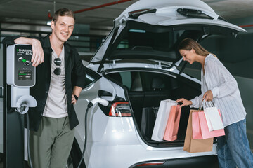 Young couple travel with EV electric car to shopping center parking lot charging in downtown city showing urban sustainability lifestyle by green clean rechargeable energy of electric vehicle innards
