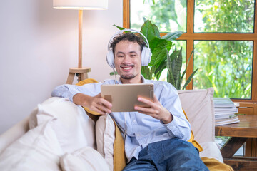 Entrepreneur businessman comfortable sitting relax lifestyle sofa couch listening music modern headphones technology tablet at indoor home apartment, adult man smiling joy fun digital tablet earphones