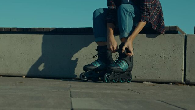Low section of active stylish female roller skater sitting on bench, putting on inline skates while preparing for rollerblading on city street at sunrise.