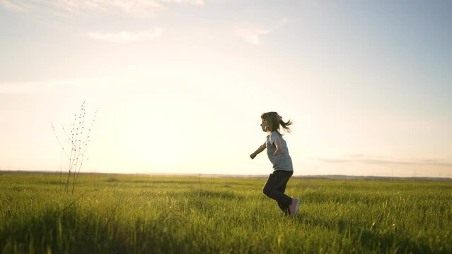 Happy child run her arms to sides. Girl play in park at sunset. Little girl run through green grass in the field. Dream girl. Cheerful child runs smiling and dreaming. Winner girl strives for success