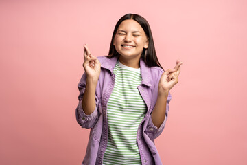 Dreaming smiling teenage girl making wish crossing fingers with closed eyes isolated on pink background. Brunette cute teen waiting expecting pleasant event, experience. Dreams desires, plans concept.